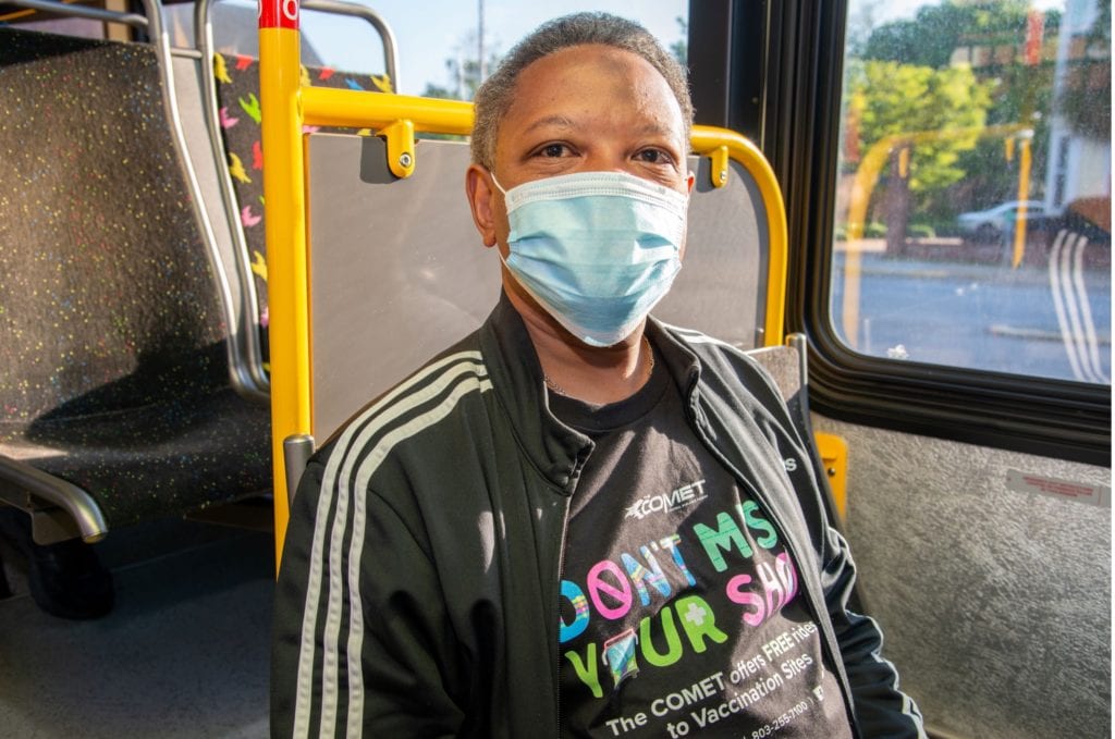 Photo of a middle-aged Black man wearing a black jacket and black t-shirt that says: ‘Don’t Miss Your Shot,’ smiling while wearing a facemask. He sits on a bus in a seat by the window.]