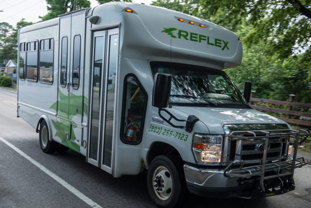 A white bus with green text that says 'Reflex' and 803-255-7123. The bus is driving down the road. 